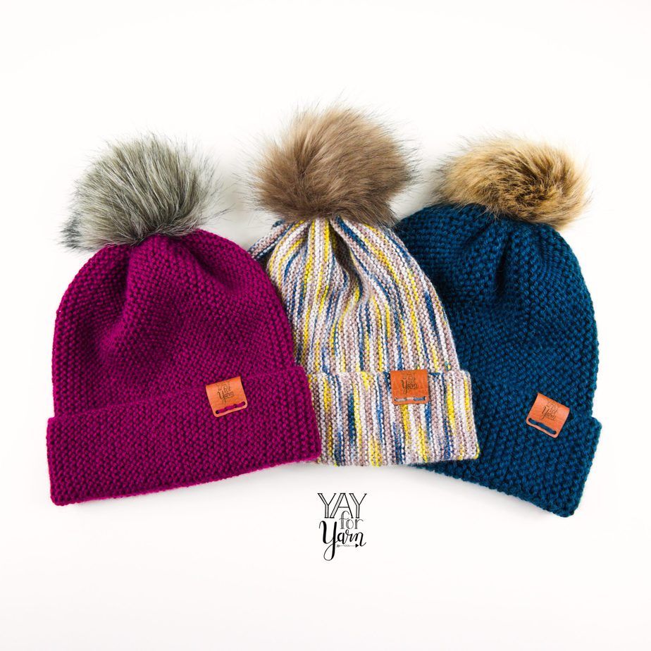 three hand knit hats with faux fur pom poms and leather labels on white background