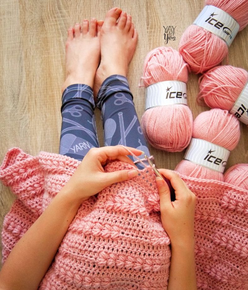 woman crocheting a pink blanket, wearing black yarn-print leggings