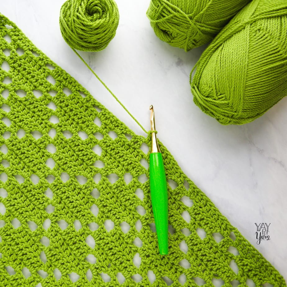 green diamond stitch crochet project with several skeins of green yarn and lime green Furls crochet hook on marble background