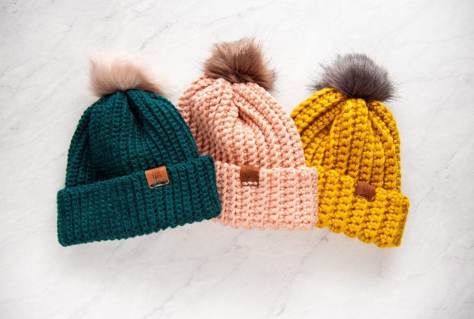 dark teal green, pale pink, and mustard yellow crochet hats with fur pom poms on white marble