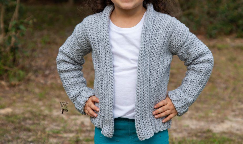 The child is outdoors wearing a gray crocheted cardigan with a white shirt and matching teal green pants and holding her waist with two hands