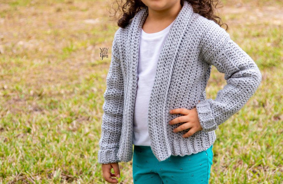 The child is outdoors wearing a gray crocheted cardigan with a white shirt and matching teal green pants and holding her waist with one hand