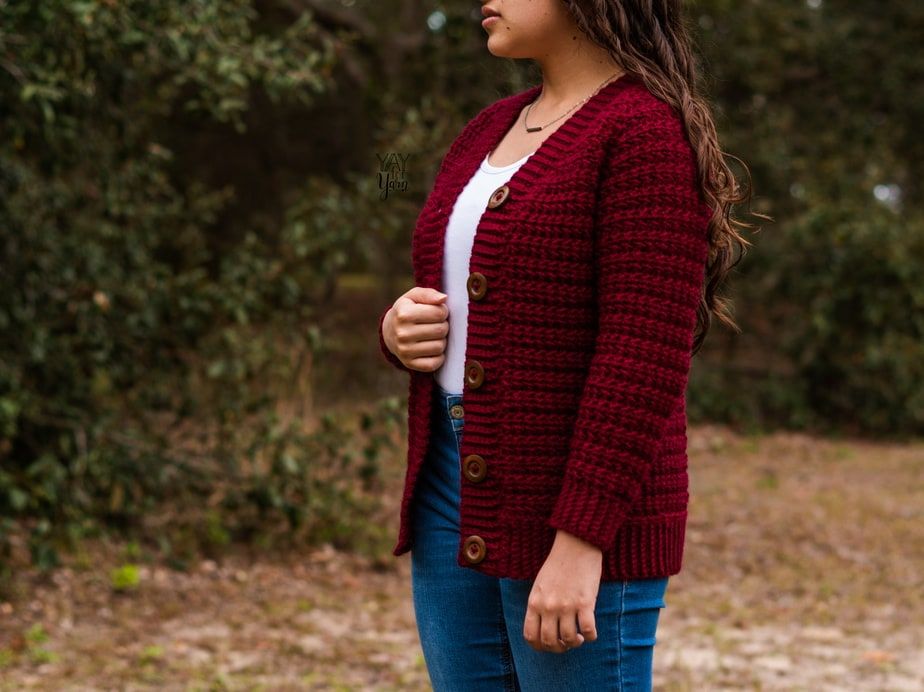 Side-view of a woman wearing crimson crochet cardigan outdoors, with white top and jeans