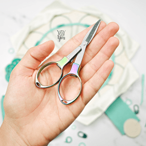 hand holding rainbow folding scissors with knitting needle set in background