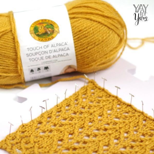 On top of an interlocking white rubber mat with over ten T needles are one yellow yarn and another with the text Lion Brand Yarns Touch of Alpaca on the side.